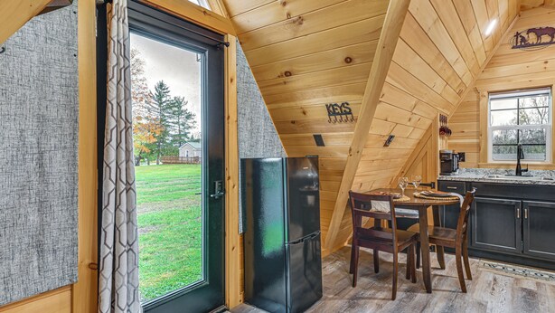 A wooden cabin interior with a dining area, a black refrigerator, and a view of the outdoors. - 5 Mi to Wine Trails: Finger Lakes Tiny Home (Ovid)