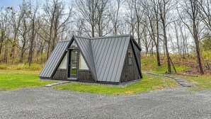 A modern, triangular-shaped cabin with a metal roof, surrounded by a gravel driveway and a grassy area. - 5 Mi to Wine Trails: Finger Lakes Tiny Home (Ovid)