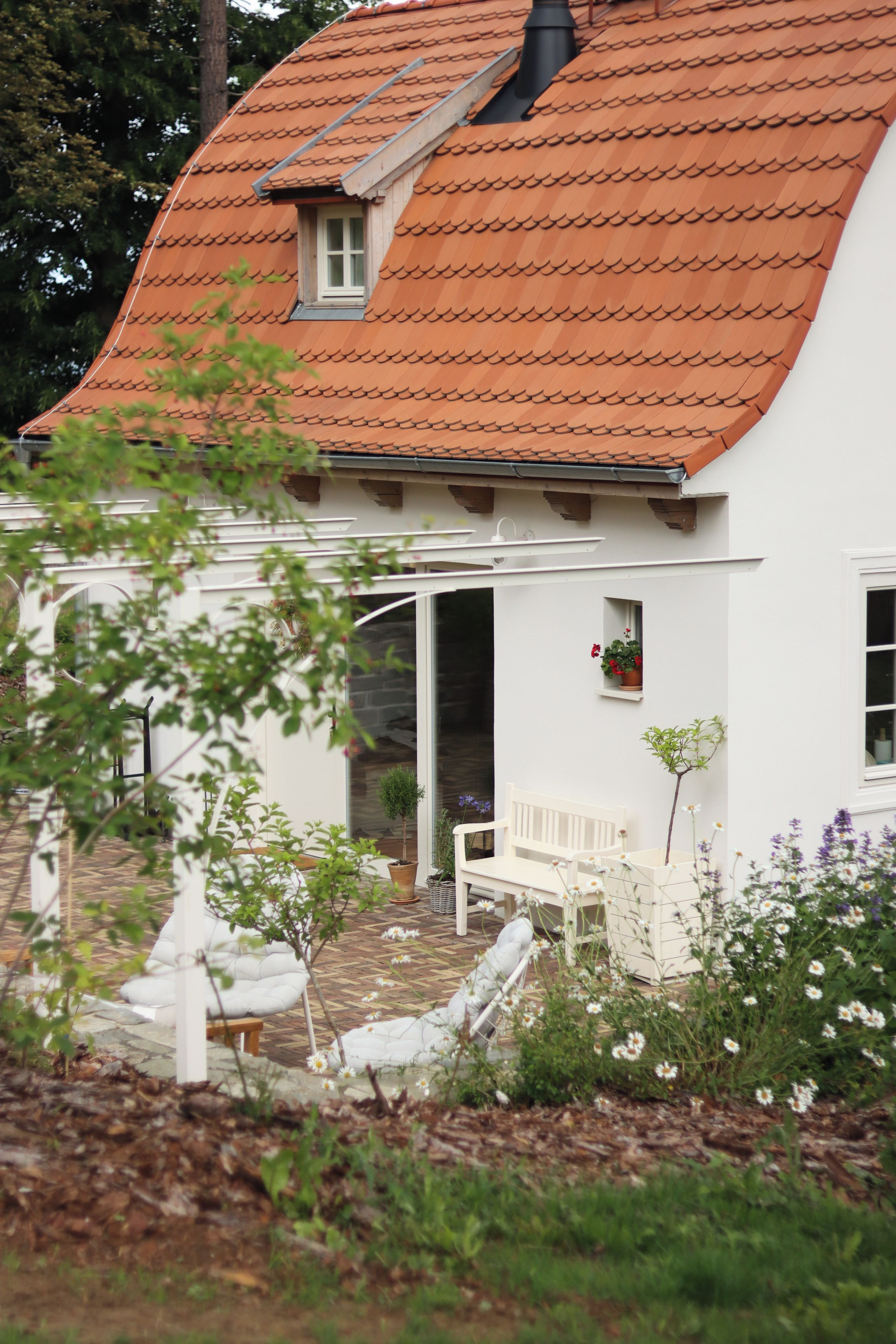 A white house with a red-tiled roof, a white bench, and a garden with various plants.