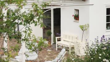 A white house with a red-tiled roof, a white bench, and a garden with various plants.