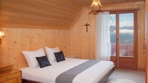 A wooden-paneled bedroom with a bed, pillows, a bedside table, a cross on the wall, and a view through a window.