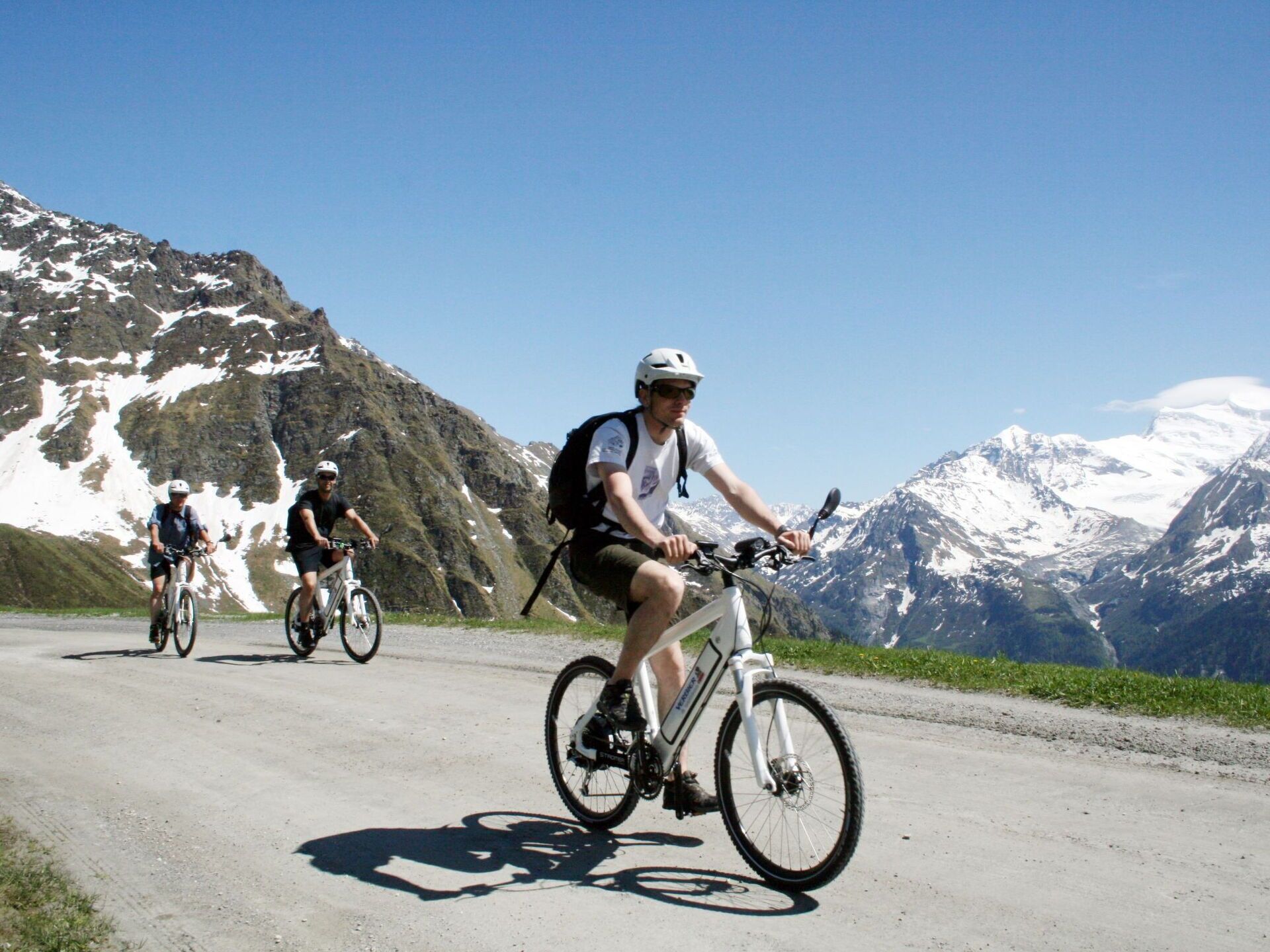 Three cyclists on a paved road with snow-capped mountains in the background.