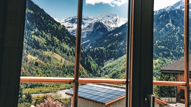 A mountain view from a window with a balcony, solar panels, and a wooden cabin. - Lovely apartment for 6 people with WIFI, balcony and panoramic view (Rabius)