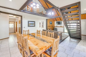 Un vaste espace dédié à la salle à manger, avec une grande table et des chaises en bois, un escalier avec des poutres apparentes et un téléviseur fixé au mur.