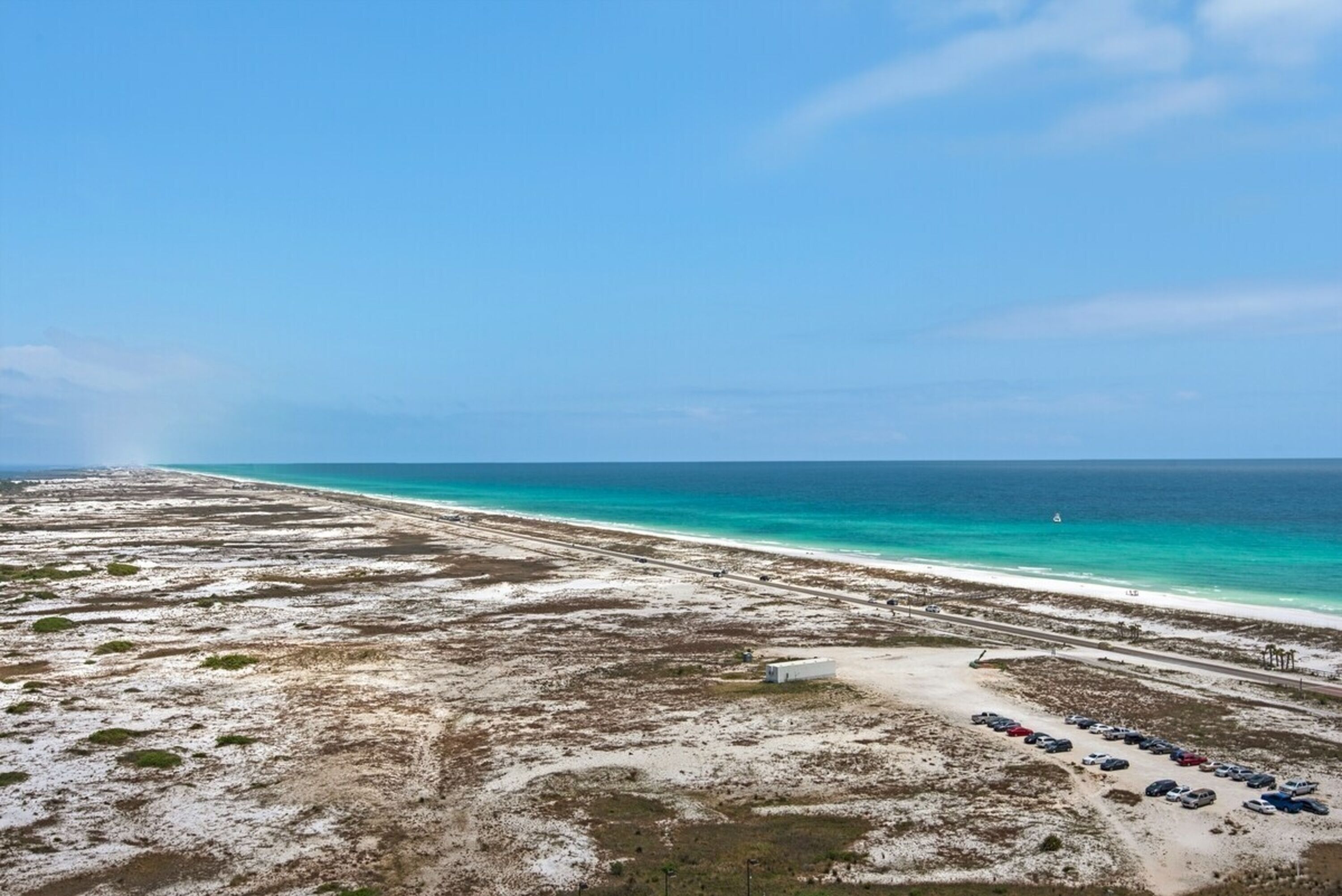 Vista aérea de una playa con cielo despejado, zona arenosa con vegetación escasa y una fila de coches aparcados a lo largo de la orilla.