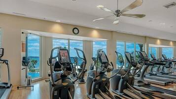 Un gimnasio con varias cintas de correr y elípticas, grandes ventanales con vista al océano y un ventilador de techo.