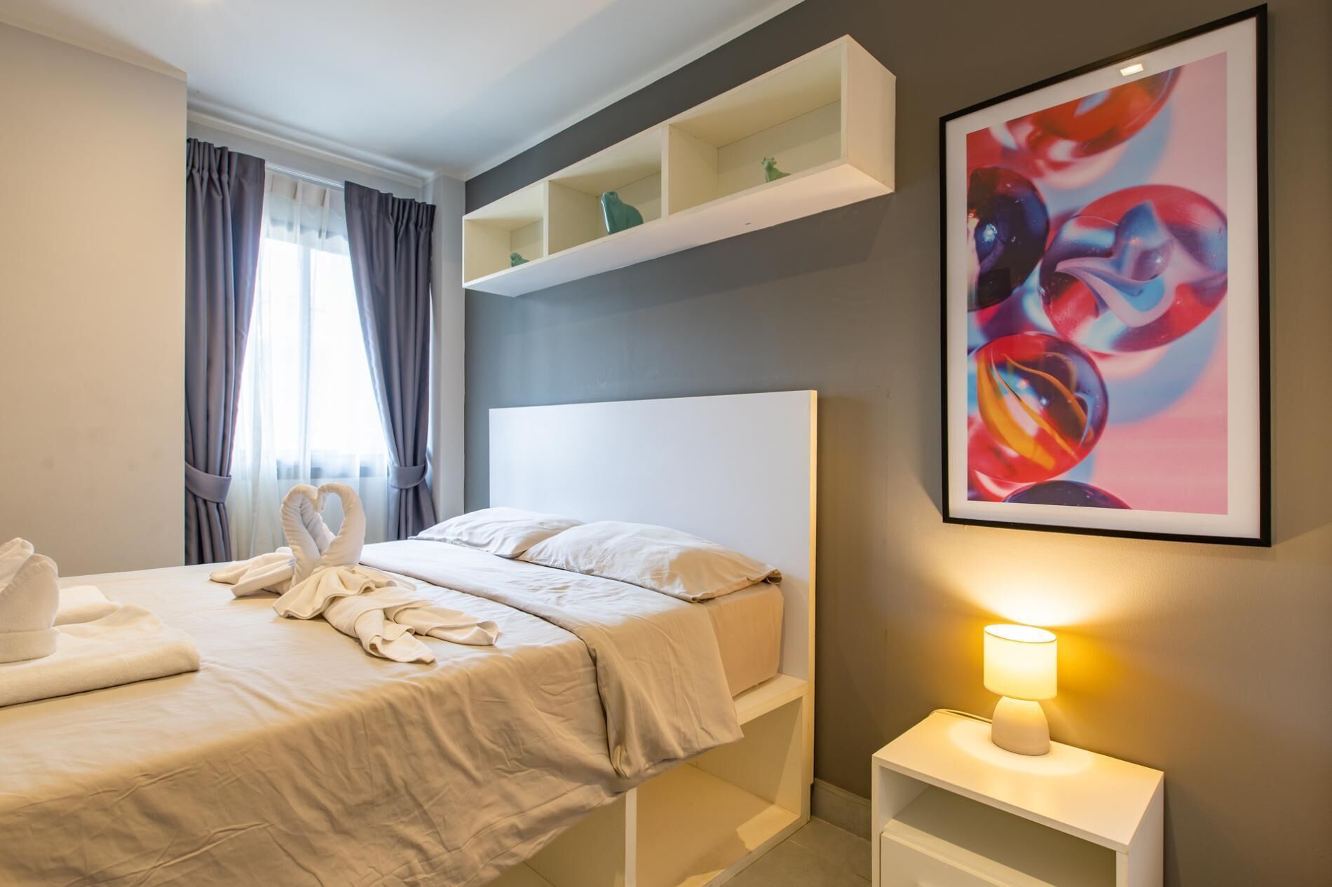 A modern hotel room with a neatly made bed, a bedside lamp, and a colorful abstract painting on the wall.