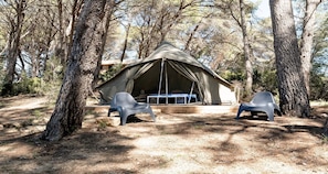 A tented accommodation with a bed and two plastic chairs in a forested area.