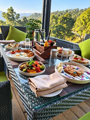A dining table set with various dishes, a glass of water, and a view of a forested landscape. - Anupraas Resort (Lansdowne)