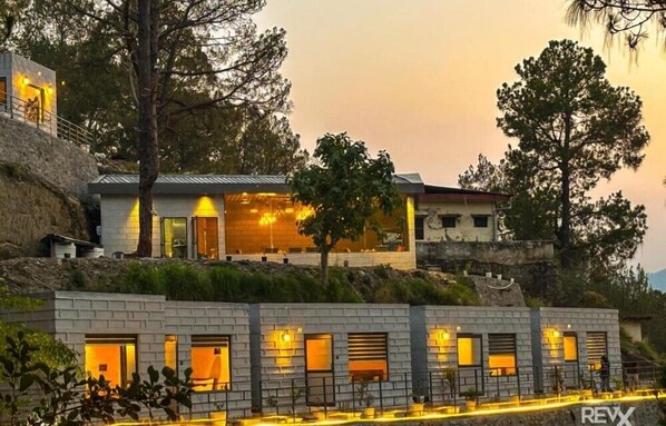 A modern building with multiple rooms, a stone retaining wall, and trees in the background. - Anupraas Resort (Lansdowne)