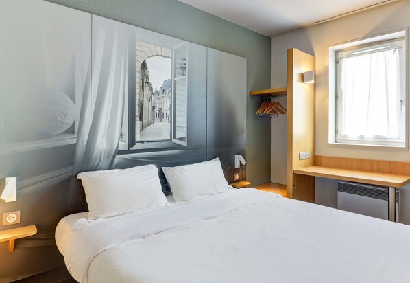 A hotel room with a large bed, a nightstand, a wall-mounted lamp, a window with curtains, and a coat rack. - B&B Hotel Dijon Les Portes Du Sud (Marsannay-la-Cote)