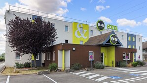 Un hotel B&B con una fachada distintiva de color amarillo y verde, un gran árbol en frente y un estacionamiento con plazas para discapacitados.