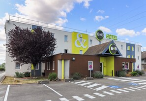 A B&B Hotel with a distinctive yellow and green facade, a large tree in front, and a parking area with disabled parking spaces. - B&B Hotel Dijon Les Portes Du Sud (Marsannay-la-Cote)