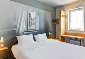 Hotellihuone, jossa on sänky, yöpöytä, seinään kiinnitetty valaisin, ikkuna ja naulakko.