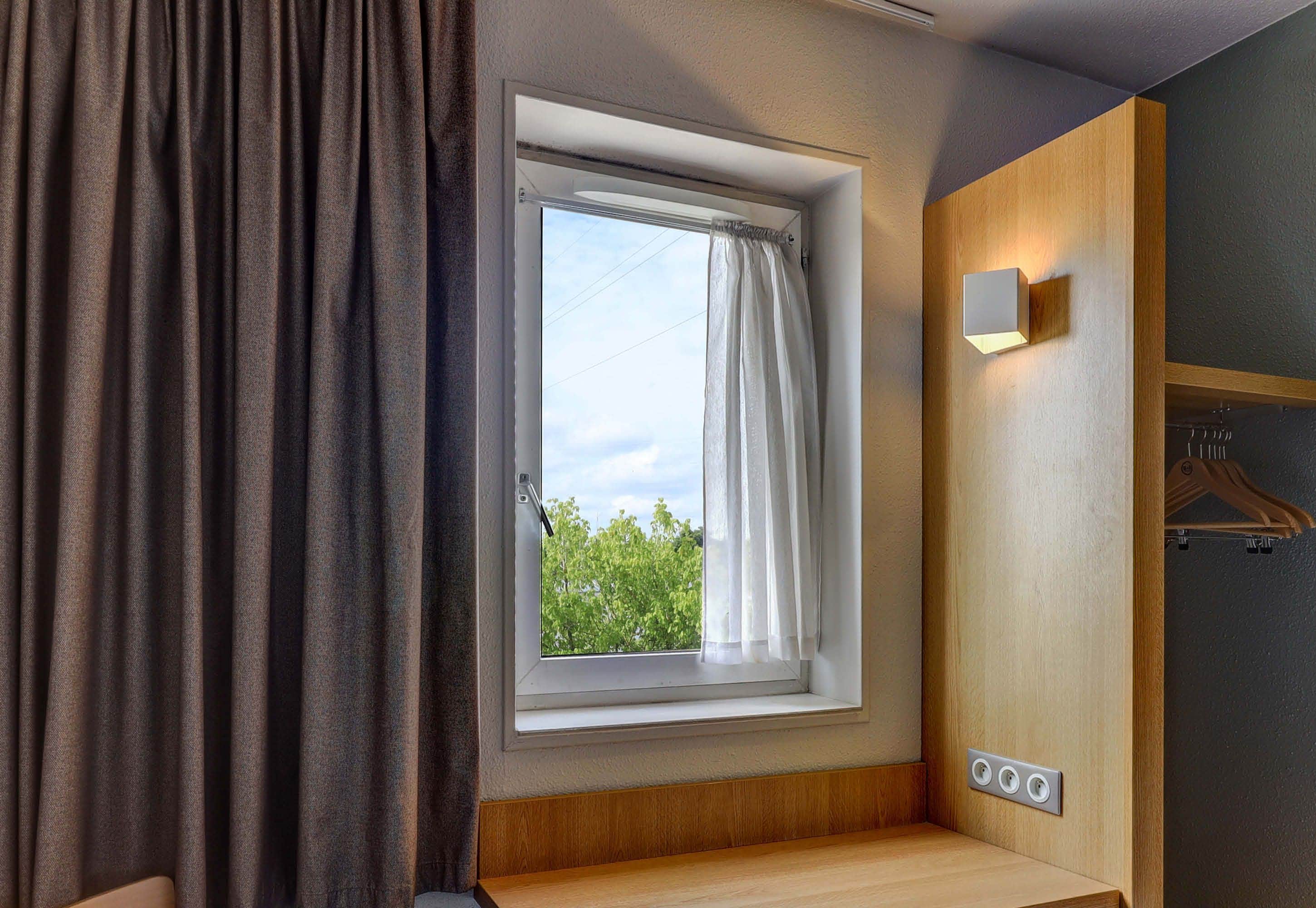 A room with a window, a curtain, a wooden bench, a wall-mounted light, and a power outlet.