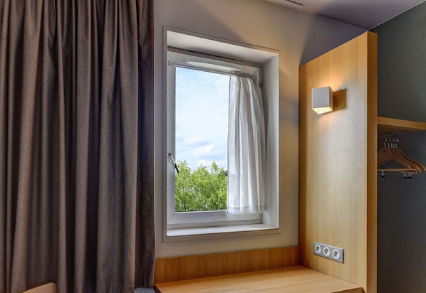 A room with a window, a curtain, a wooden bench, a wall-mounted light, and a power outlet. - B&B Hotel Dijon Les Portes Du Sud (Marsannay-la-Cote)