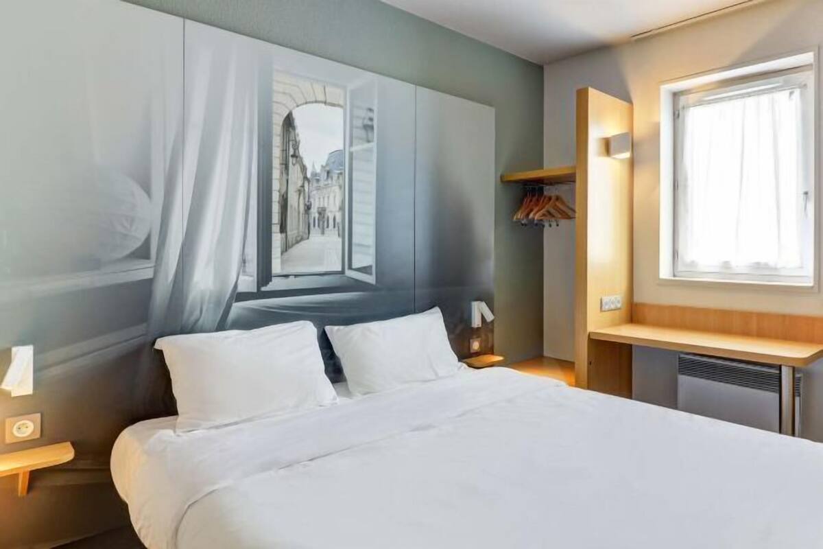 Une chambre d’hôtel avec un lit, une table de chevet, une fenêtre, une étagère fixée au mur avec des vêtements et un tableau représentant une scène de rue.