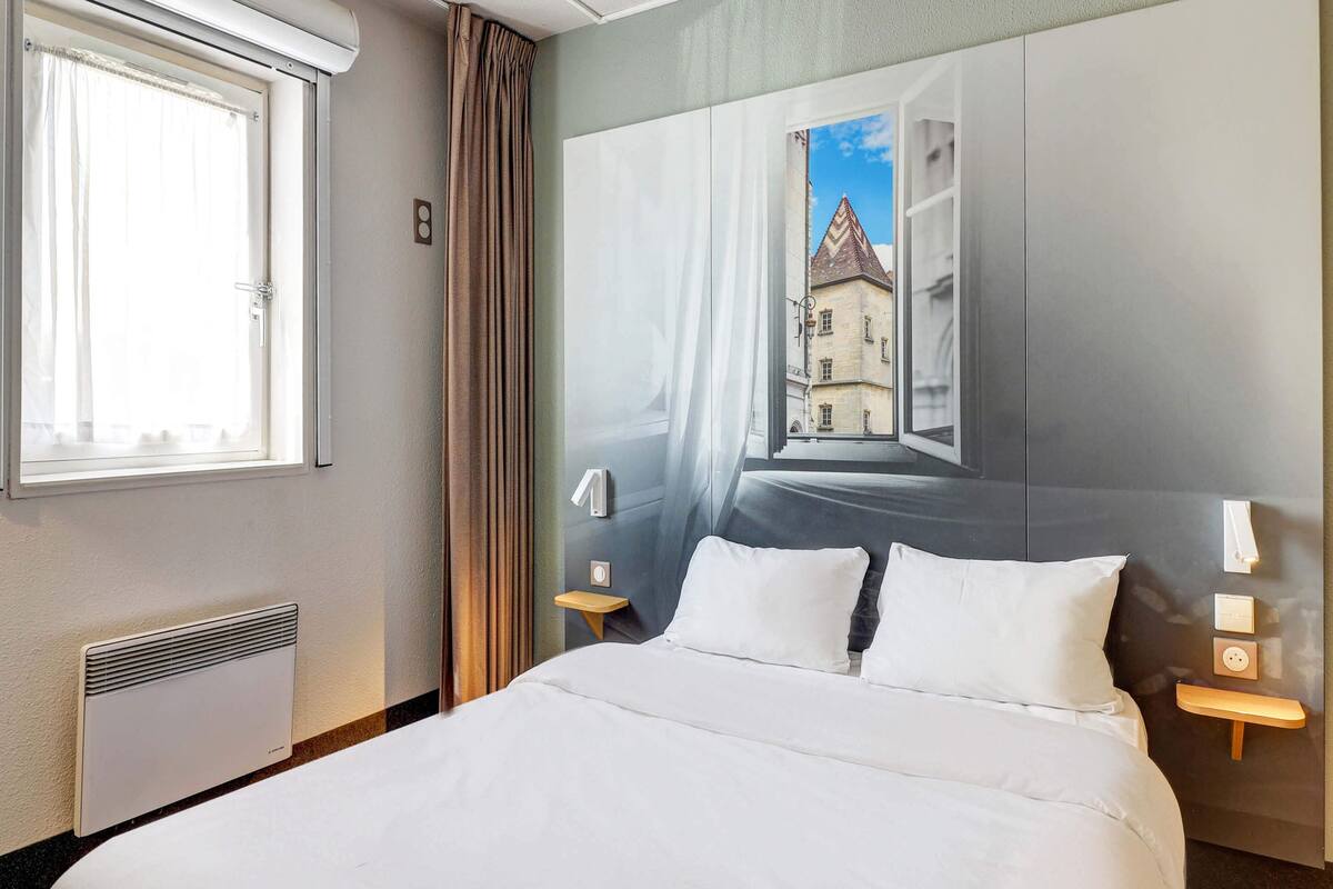 Une chambre d’hôtel avec un grand lit, deux tables de chevet, une fenêtre avec des rideaux, un radiateur et un tableau représentant un bâtiment accroché au mur.