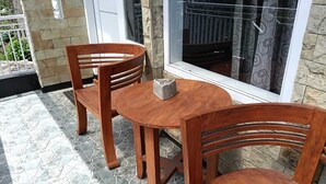 Een terras met een ronde houten tafel en twee stoelen, een vloer van stenen tegels en een raam met uitzicht op een balkon.