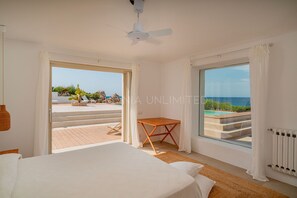 Une chambre à coucher avec un lit, une petite table en bois et une vue sur la mer grâce à de grandes fenêtres.