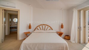 A bedroom with a bed, two bedside tables, a ceiling fan, and a view of a bathroom. - Villa Janas Eterea - Sardinia Unlimited (Portobello Di Gallura)