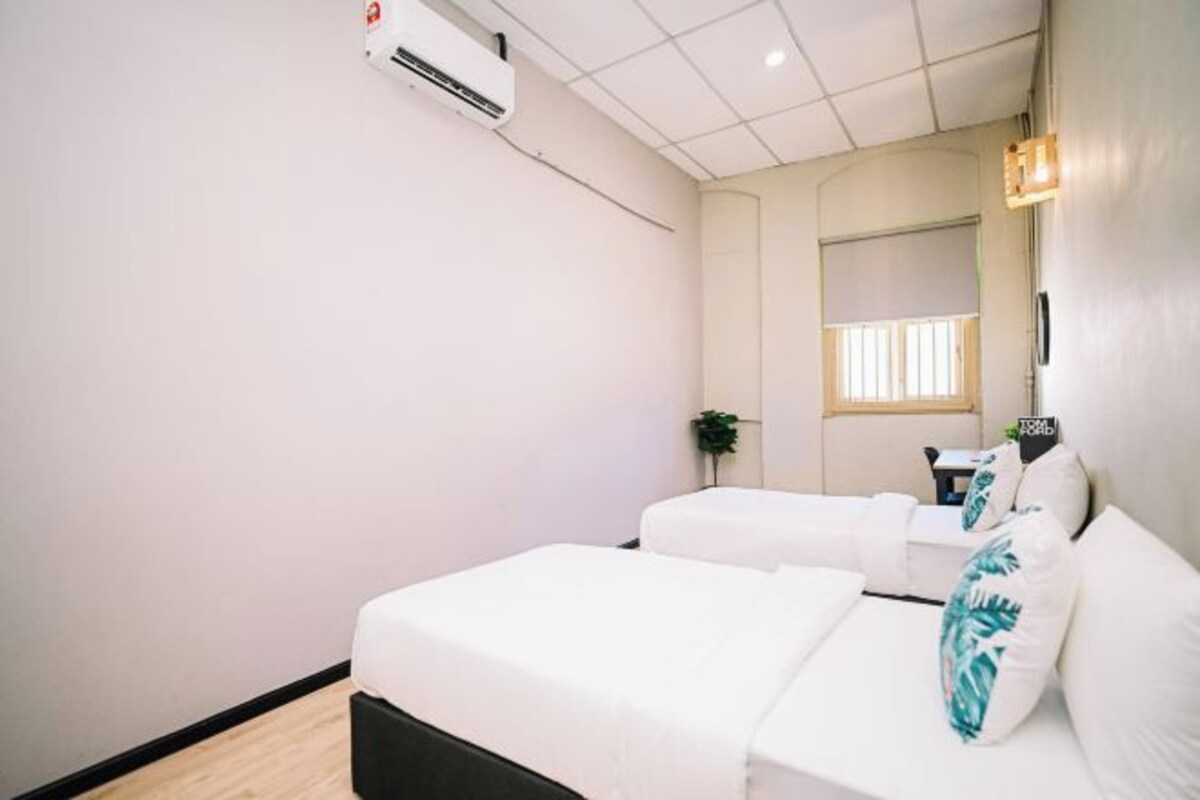 Ein Hotelzimmer mit zwei Betten, einem kleinen Tisch, einem Stuhl, einer Klimaanlage, einem Fenster mit Jalousien und einer Wandleuchte.