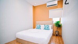 Un lit bien fait dans une pièce au plancher en bois, avec un climatiseur fixé au mur et une plante en pot placée à côté du lit.