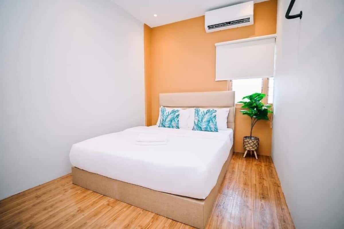 A neatly made bed in a room with wooden flooring, an air conditioning unit on the wall, and a potted plant beside the bed.