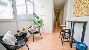 Un couloir avec un plancher en bois, des chaises noires avec des coussins, une petite table et une poubelle bleue.