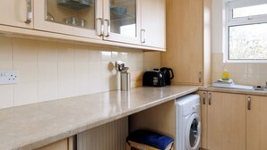 A compact kitchen with wooden cabinets, a built-in washing machine, and a small stool. - Spacious 2-Bedroom Flat Near Rottingdean Beach & Windmill (Brighton and Hove)