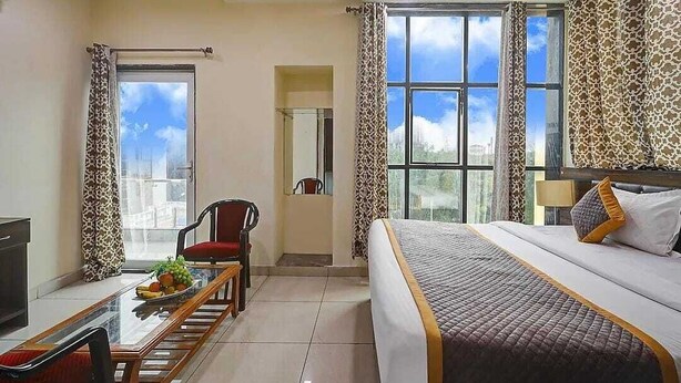 A hotel room with a bed, a desk with a chair, a dining table with fruit, and large windows with curtains. - Hotel Vibe Blue (Noida)
