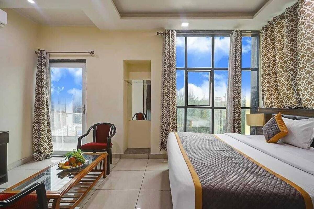 A hotel room with a bed, a desk with a chair, a dining table with fruit, and large windows with curtains.
