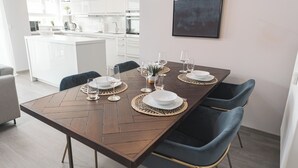 A modern dining area with a wooden table set for four, surrounded by dark blue chairs. A kitchen with white cabinets is visible in the background.