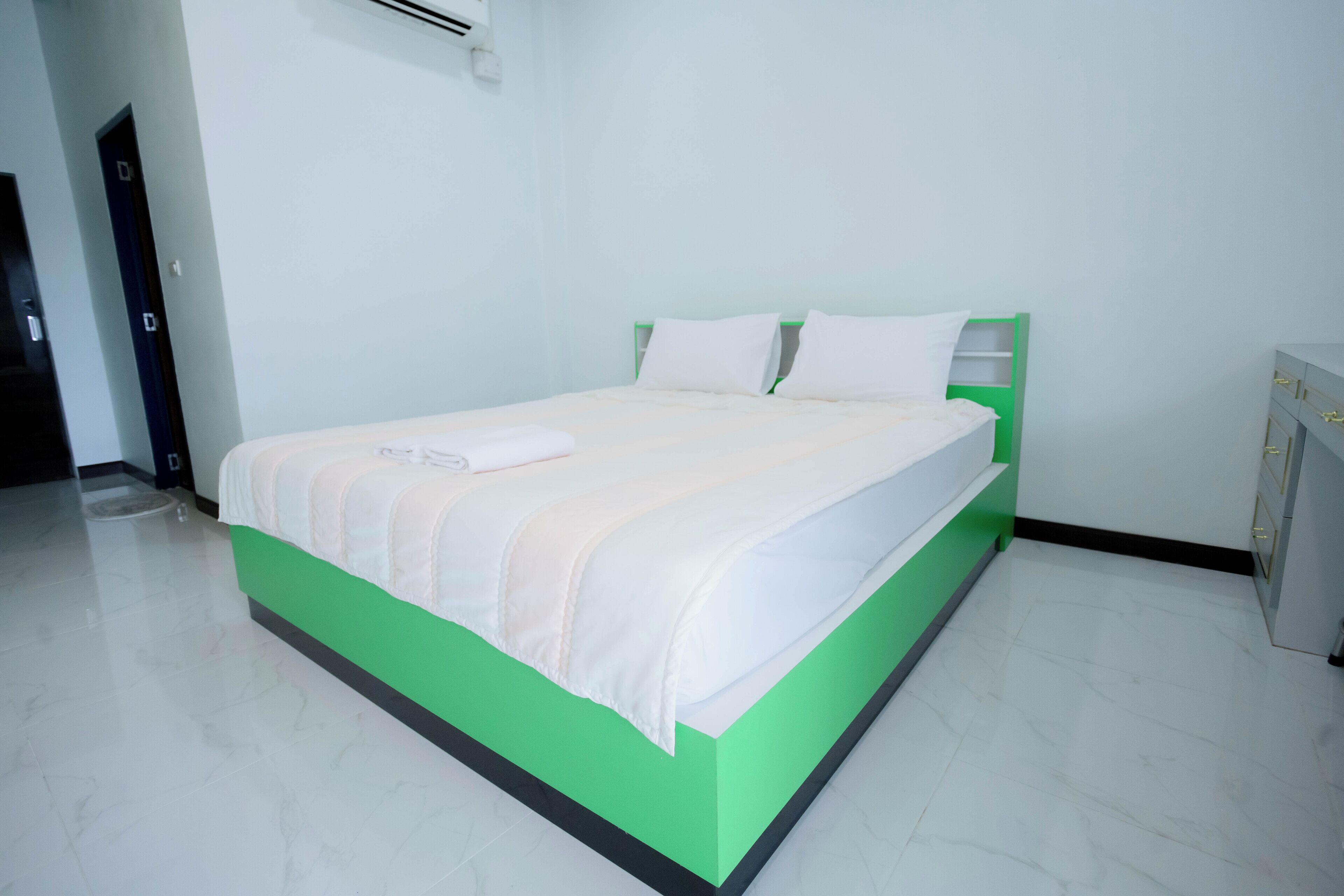 Una cama bien hecha con estructura verde y ropa de cama blanca en una habitación con paredes blancas y suelo de baldosas.