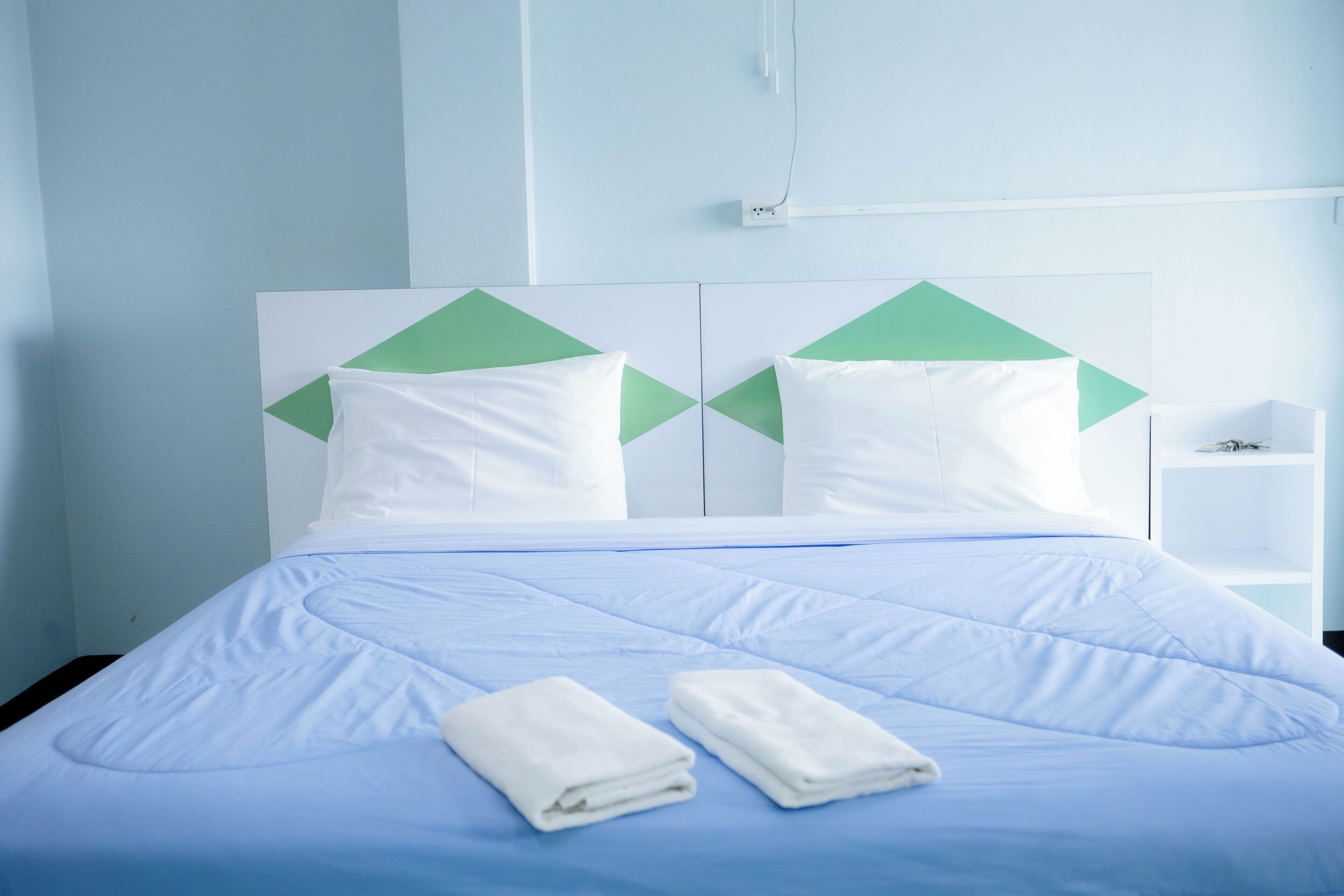 A neatly made bed with a blue bedspread and two white pillows, a white bedside table, and a wall with geometric patterns.