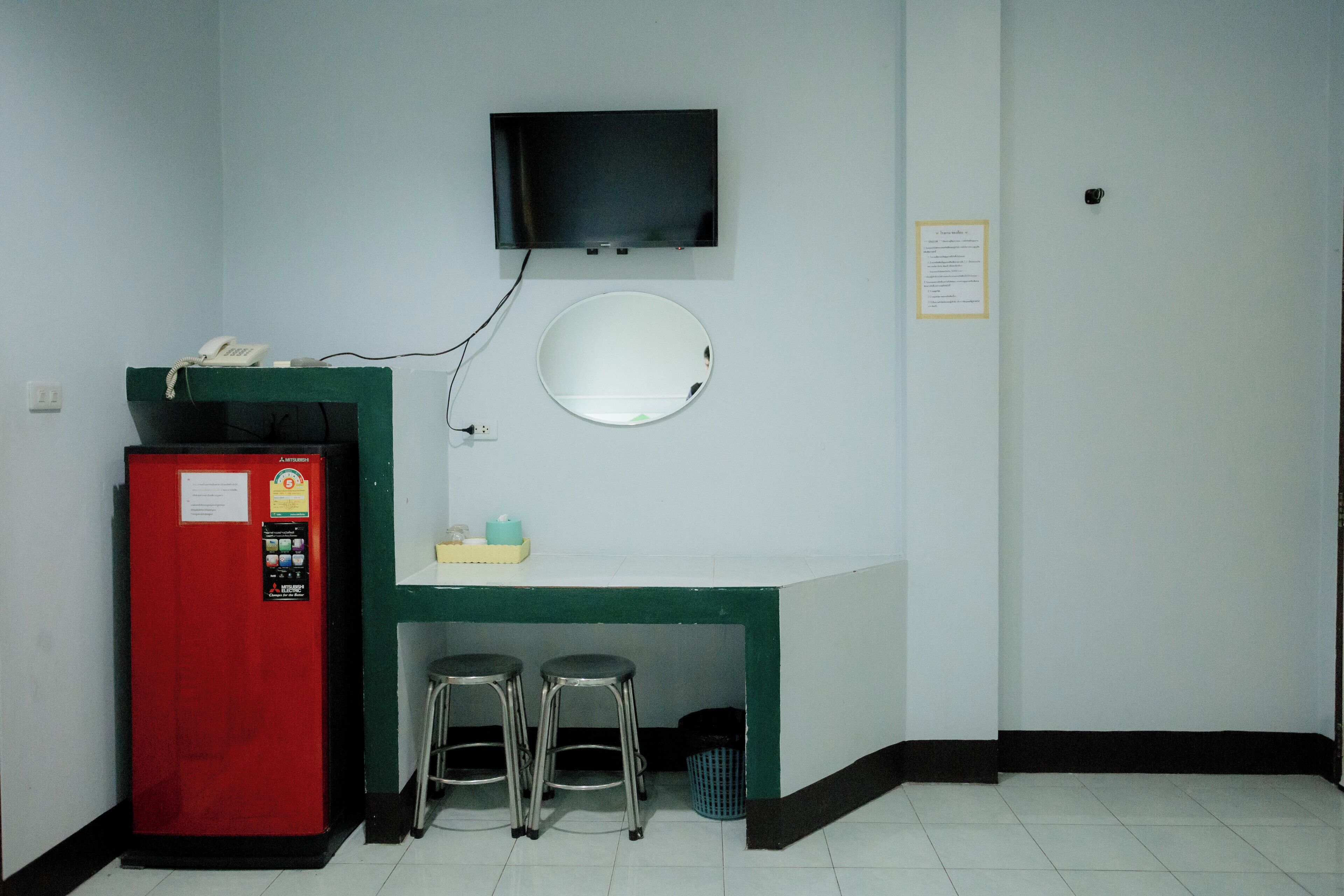 A small room with a red vending machine, a TV mounted on the wall, a round mirror, and two stools.
