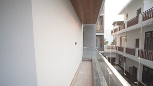 Un balcón estrecho con barandilla de vidrio, techo de madera y vista a un edificio de varios pisos con balcones.