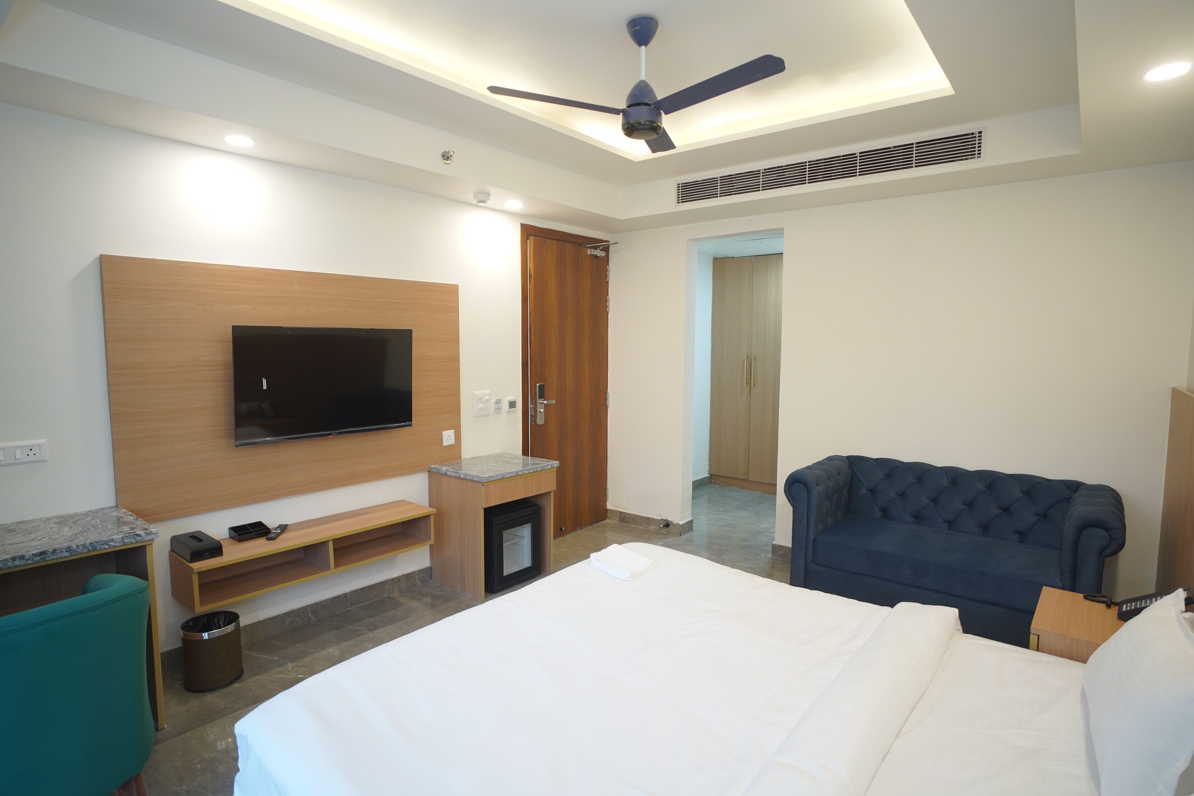 A modern hotel room with a bed, a TV mounted on the wall, a ceiling fan, and a dark blue sofa.