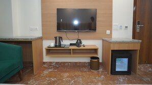 Una habitación moderna con un televisor de pantalla plana montado en la pared, un pequeño estante de madera con una tetera y una bolsa, un refrigerador compacto y un cubo de basura.