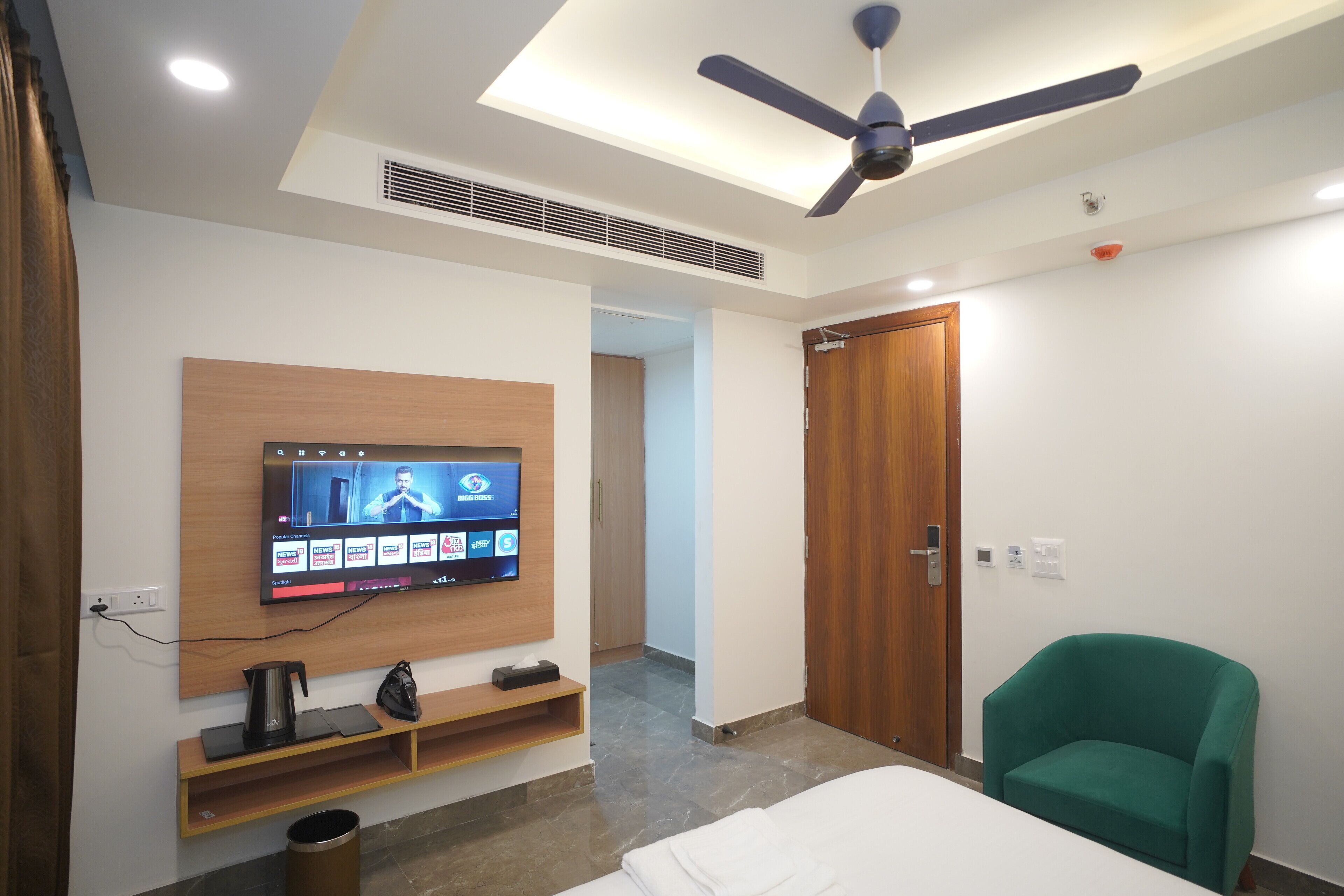 A modern hotel room with a flat-screen TV mounted on the wall, a ceiling fan, a wooden door, and a green armchair.