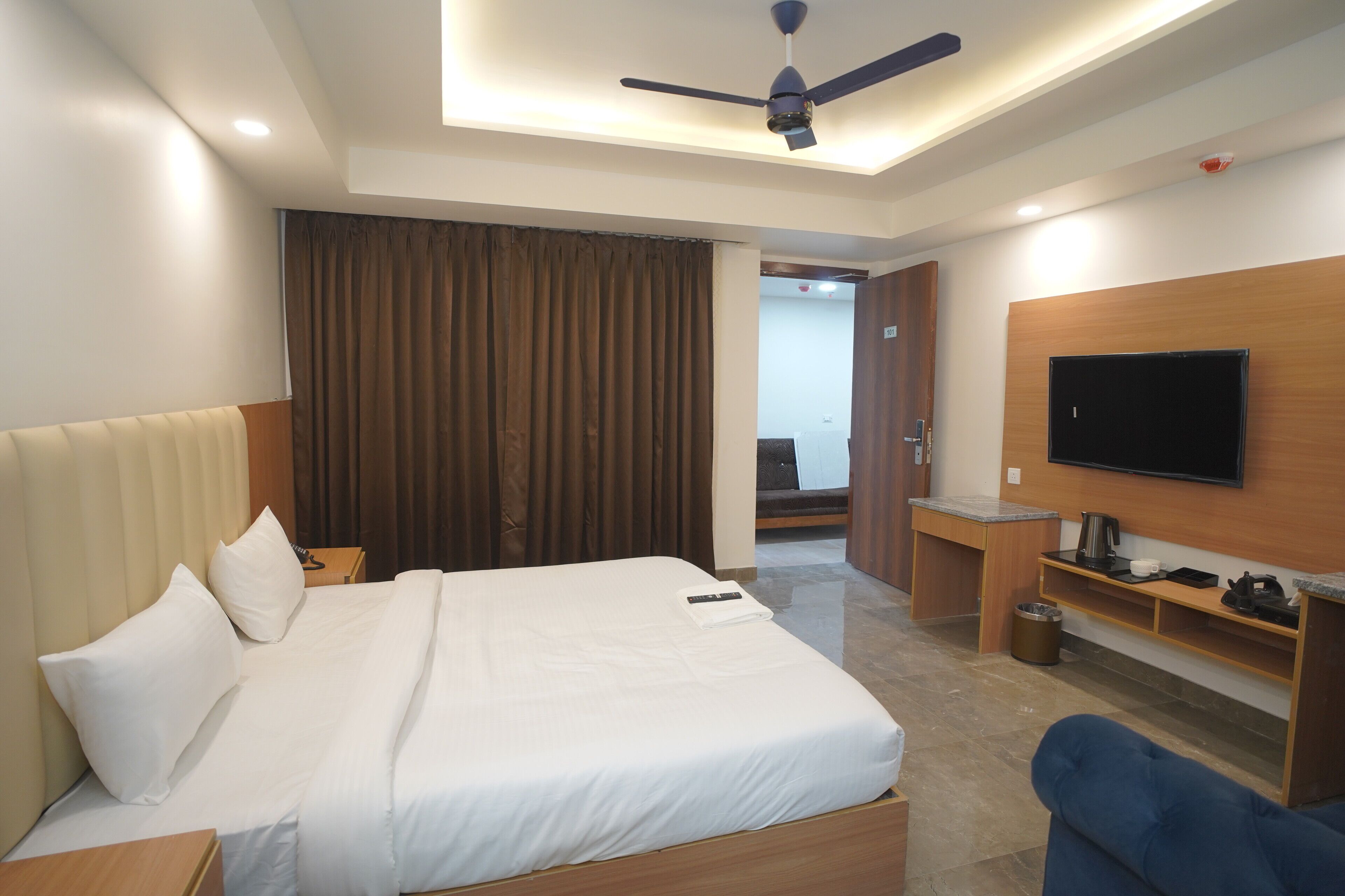 A hotel room with a bed, a ceiling fan, a TV mounted on the wall, and a small kitchenette area.