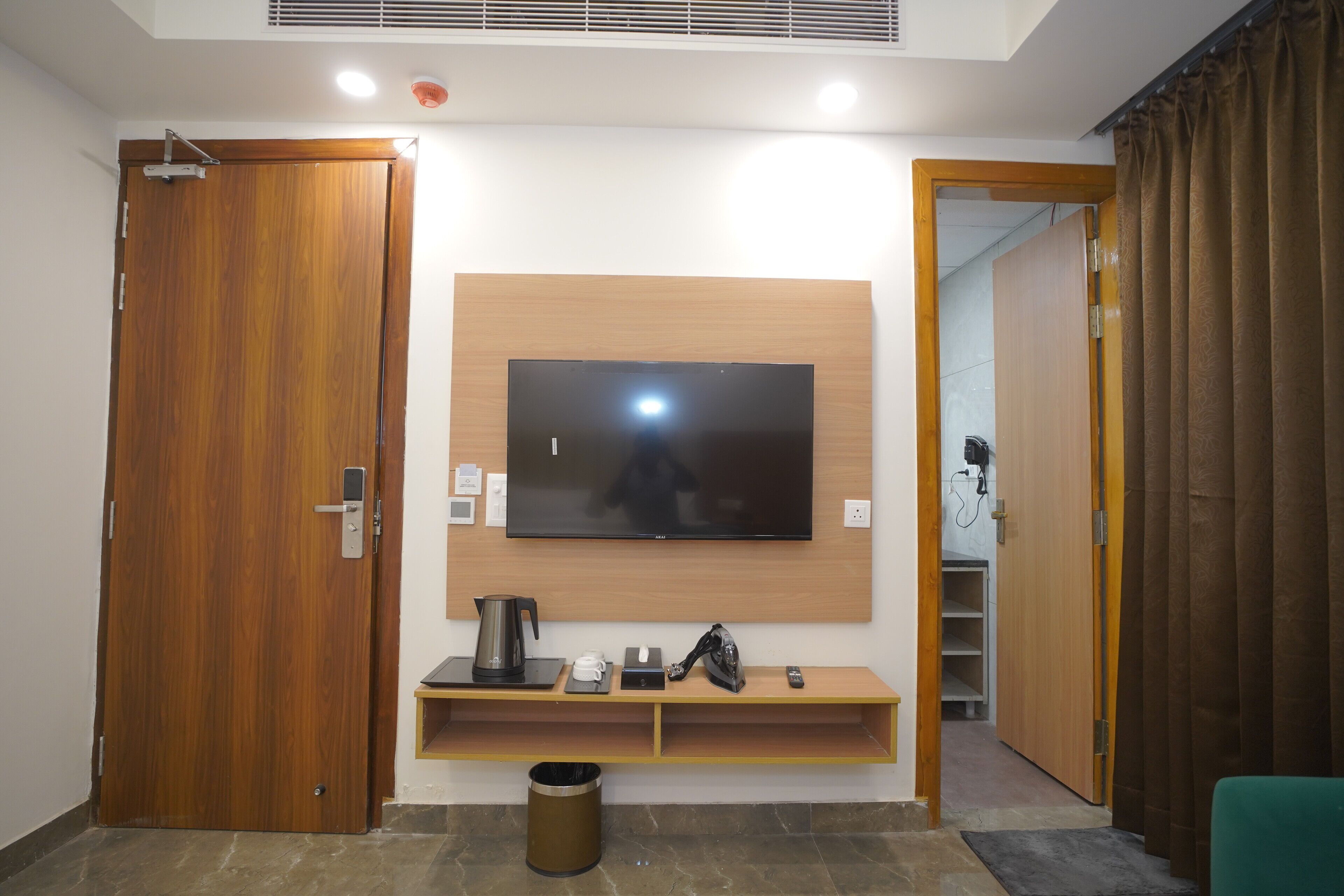 Une chambre d’hôtel avec une porte en bois, un téléviseur fixé au mur, une machine à café, une bouilloire, une poubelle et une chaise verte.