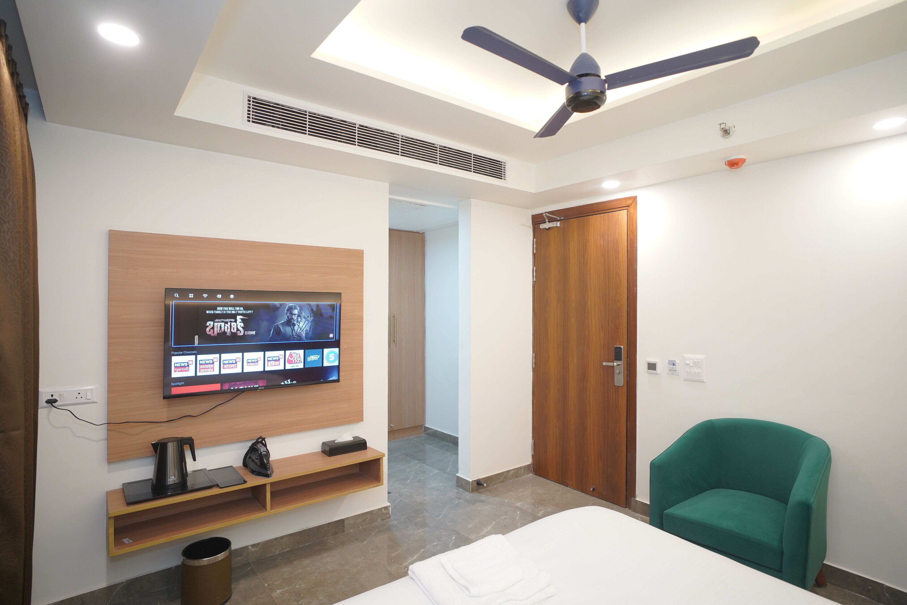 Una camera d'albergo moderna con una TV a schermo piatto montata a parete, una ventola a soffitto, un armadio in legno, una poltrona verde e un letto rifatto con cura.