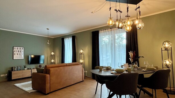 Ein großzügiger Wohnbereich mit Sofa, Esstisch und Stühlen. An der Wand ist ein Flachbildfernseher angebracht, und über dem Essbereich hängt eine Deckenleuchte.