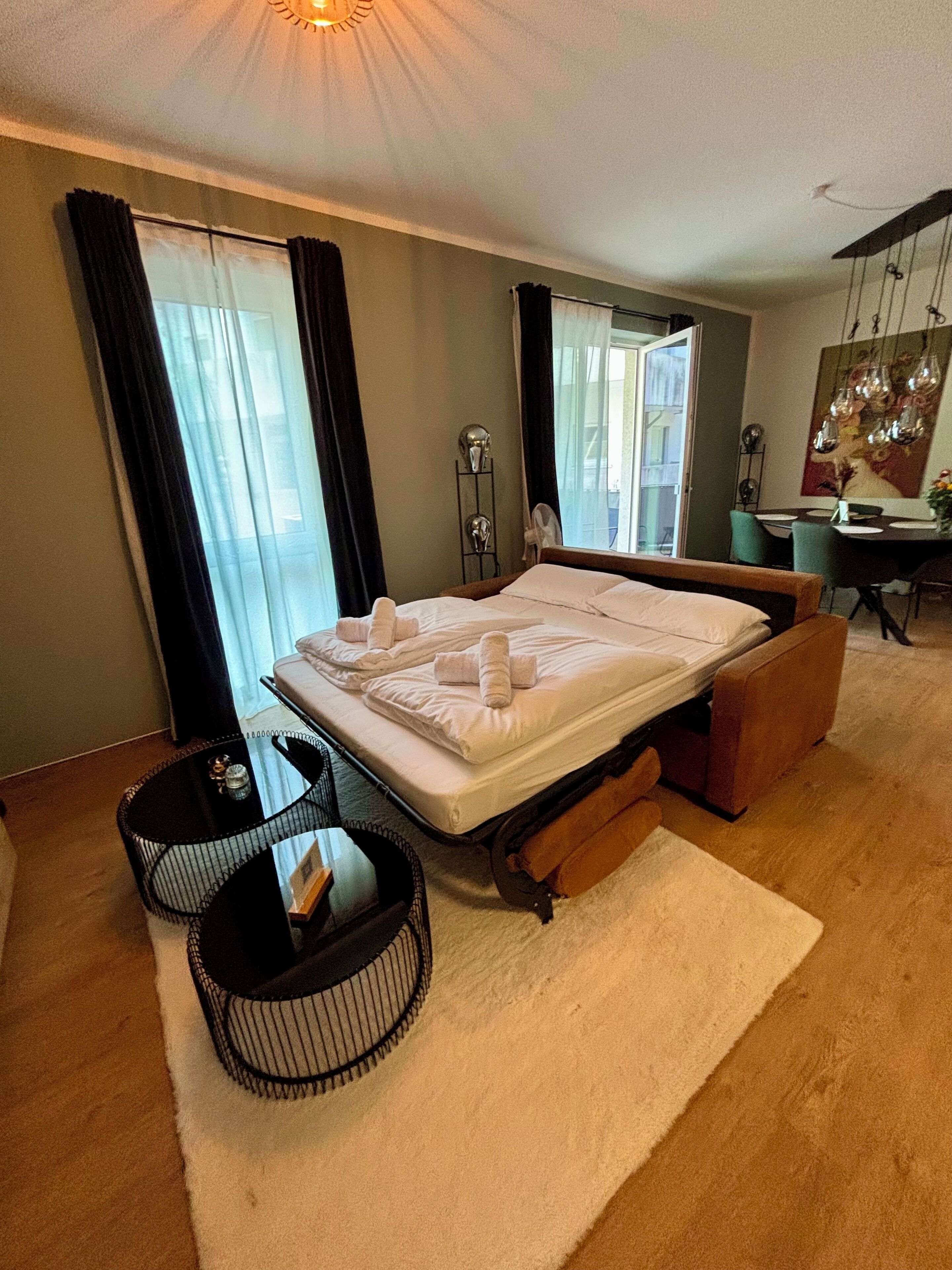 A modern bedroom with a bed, bedside tables, a dining area, and a large window with curtains.
