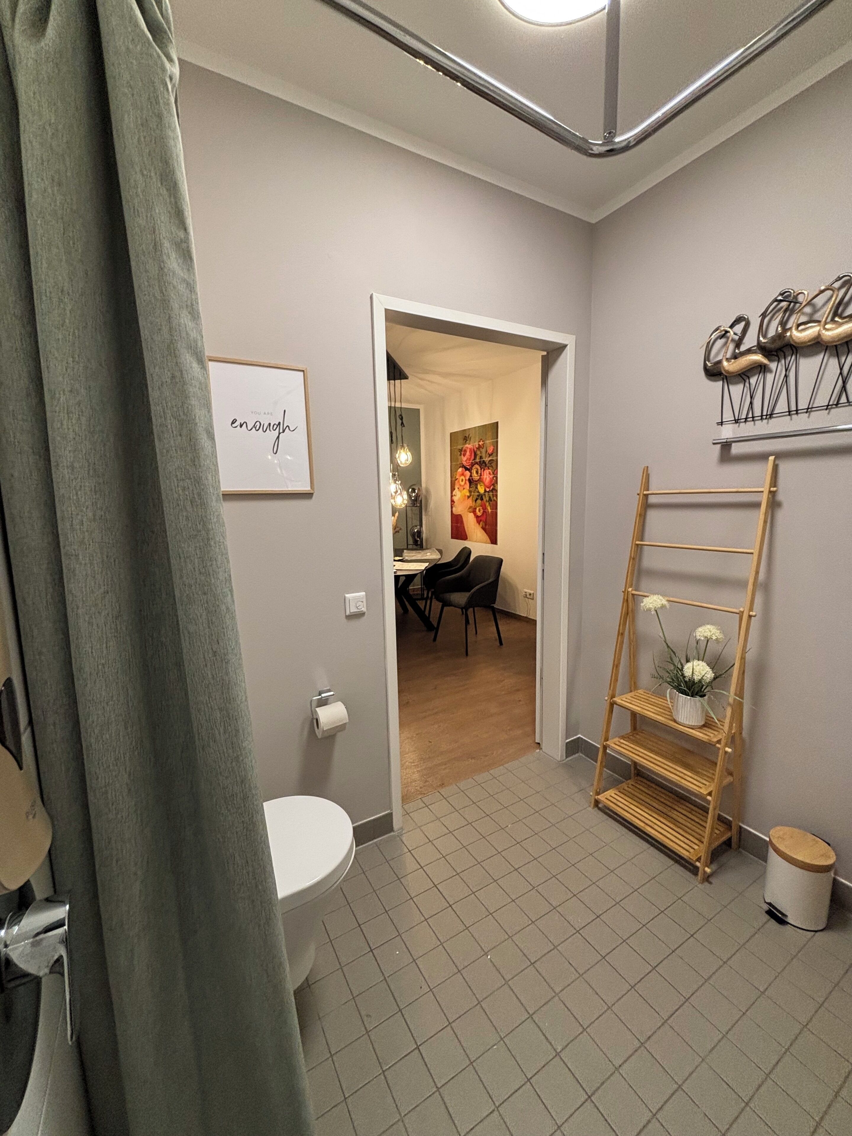 Ein Badezimmer mit Toilette, Waschbecken, einer Duschkabine, einem gerahmten Motivationszitat, einer Holzleiter-Regal mit Blumen und einer Tür zu einem Essbereich.