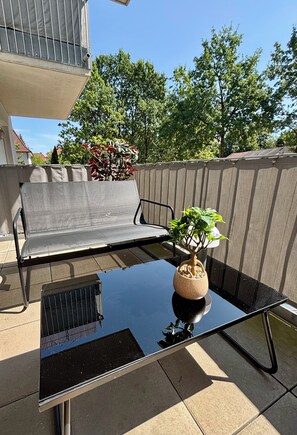 Een glazen tafel met een potplant, een grijze buitenbank en een metalen barbecue.