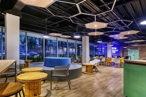 Un espacio interior moderno con mesas de madera redondas, una zona de estar azul y ventanas amplias que ofrecen vistas a zonas verdes.