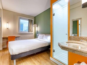Une chambre d’hôtel comprenant un lit, un bureau, une chaise, une fenêtre avec des rideaux, une salle de bain avec un miroir et un lavabo.