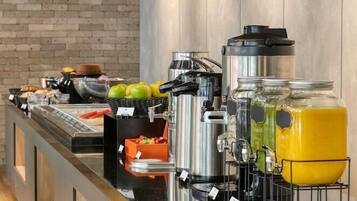 Zona de buffet con diversas estaciones de comida, que incluyen una máquina de café, fruta y dispensadores de jugo.